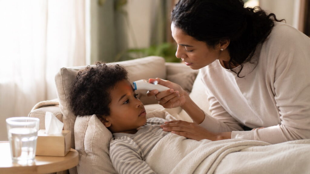Mother checking her child's forehead temperature with a digital thermometer while the child rests on a couch at home