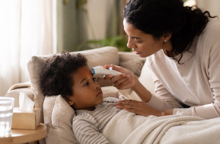 Mother checking her child's forehead temperature with a digital thermometer while the child rests on a couch at home